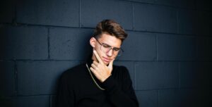 A young man in glasses and a black jumper leaning against a dark wall with his hand on his chin