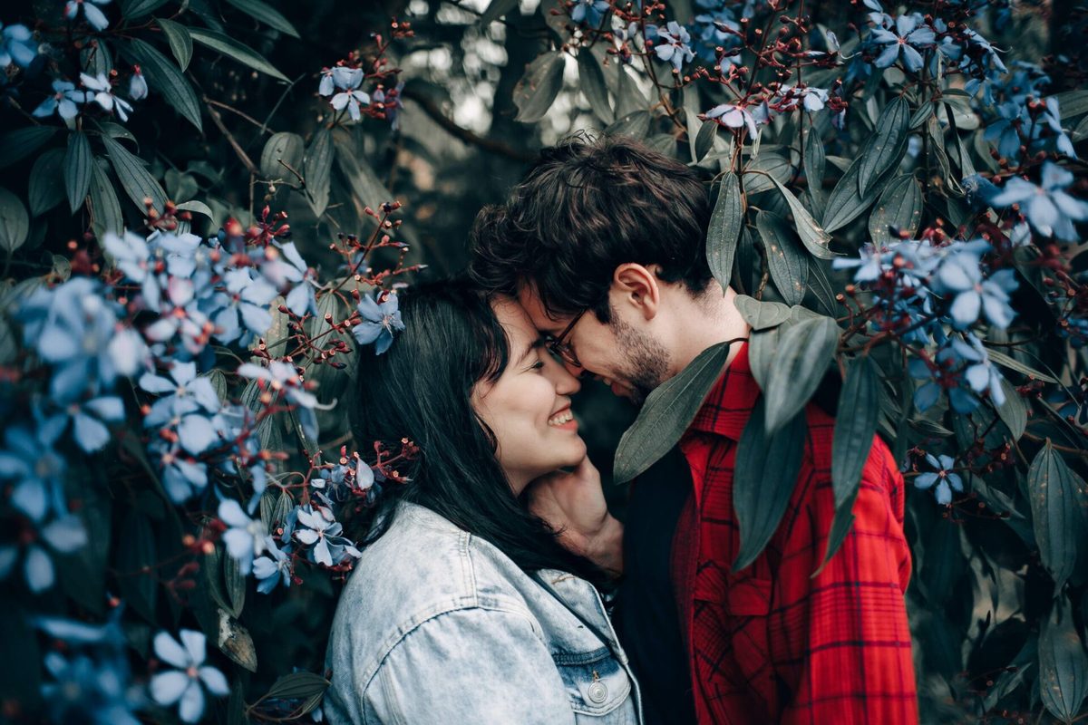 Pareja sonriente con las frentes juntas delante de un arbusto de flores azules