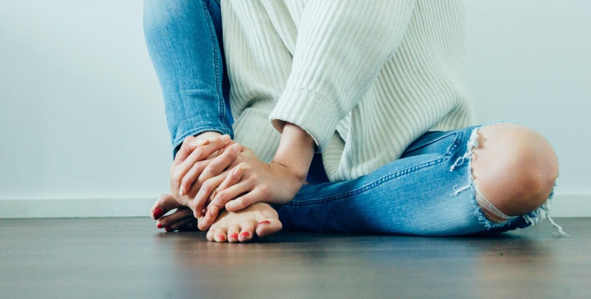 Mujer sentada en el suelo con jersey blanco y vaqueros, sujetándose los pies