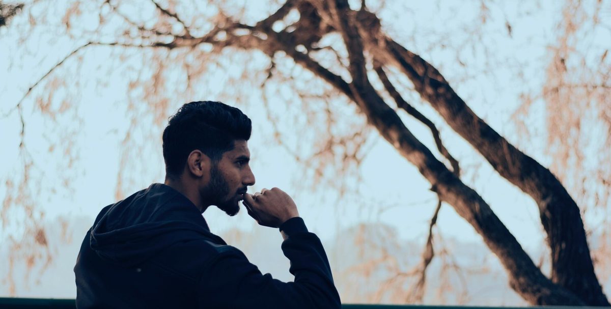 Un hombre con sudadera oscura mira pensativo bajo un árbol